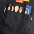 A black poly-cotton canvas apron with a pocket containing utensils and a towel, featuring a logo.