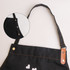 A black poly-cotton canvas full bib apron featuring a neck strap and metal buttons, with a close-up inset.