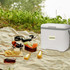 A white cooler box with a grey handle sits on a picnic blanket, surrounded by food and drinks. There is a logo on the box.