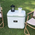 A white and green 22L cooler box sits on grass, accompanied by a lantern and a drink cup, near folding chairs.