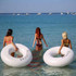 Three women wade into the water holding large inflatable swim rings, one labelled "BRUNETTE" and the other "BLONDE," against a blue sea.