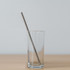 A drinking glass containing a stainless steel straw, placed on a wooden surface against a light background.