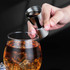 A stainless steel double jigger is held above a glass filled with iced brown liquid. The background is dark.