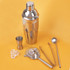 A cocktail shaker set in silver, including a shaker, jigger, strainer, tongs, and ladle on a yellow background.