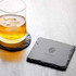 A glass of whisky sits on a round slate coaster next to a square slate coaster, both in dark grey tones.