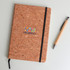 A cork notebook with a black elastic band and a pen next to it, featuring colourful branding on the cover.