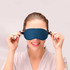 A satin sleep mask in navy blue, being held up by a smiling woman in a light-coloured robe.