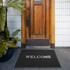 A black floor mat with a logo in front of a wooden door, surrounded by greenery.