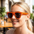 A pair of vibrant orange folding sunglasses with dark lenses, displayed on a smiling woman outdoors.