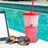 A pink and white colour changing cup with a red lid and straw, placed beside sunglasses and an e-reader by a pool.