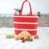 A red insulated cooler bag with white stripes, surrounded by bread, fruit, and a container, on a sandy beach.