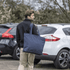 A man in a black jacket carries a large navy blue tote bag while walking past parked cars. The bag has a logo.