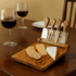 A wooden cheeseboard set featuring four stainless steel knives, cheese, and crackers, with two glasses of red wine in the background.