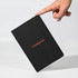 A black A5 notebook with a textured cover and an elastic band, being held by a hand. It features a logo.
