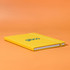 A yellow A5 notebook with an elastic band closure and a logo on the front, set against an orange background.