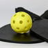 A yellow pickleball with holes, resting on a black paddle and a round black surface, featuring a logo.