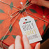 A gift box wrapped in vibrant red wrapping paper with floral patterns, featuring a white swing tag attached.