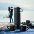 A drink bottle and two black mugs on a blue surface, with a scenic backdrop. Contains a logo.