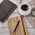 A5 wood look notebook in natural wood tones, a black notebook, a pen, coffee cup, glasses, and a magazine on a table.