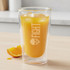 A double wall glass tumbler with a yellow drink and ice, featuring a logo, on a wooden surface.