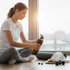 A drink bottle in black sits beside a woman preparing for a workout on a wooden floor, with dumbbells and a yoga mat.