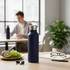 A drink bottle in navy blue with a silver lid, set on a table alongside fruit, glasses, and stationery.