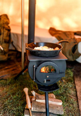 Petromax Loki2 portable tent oven and heater – wood-burning camp stove with cooking surface, foldable legs, and stovepipe, shown in use cooking with a pan.