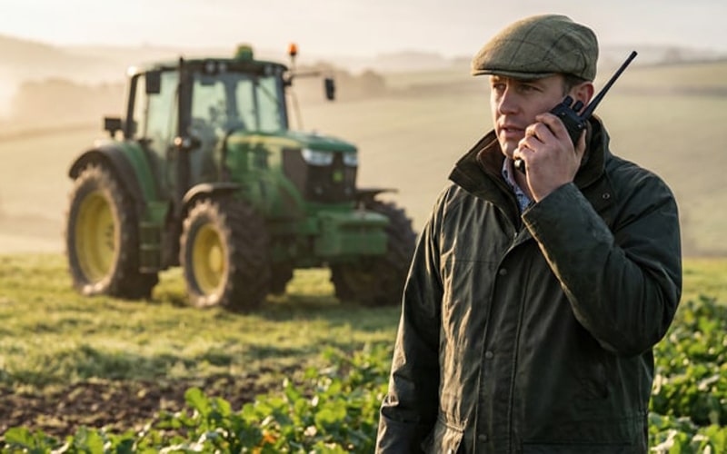 A UK farmer using a rugged two-way radio next to a tractor in a wide agricultural field.