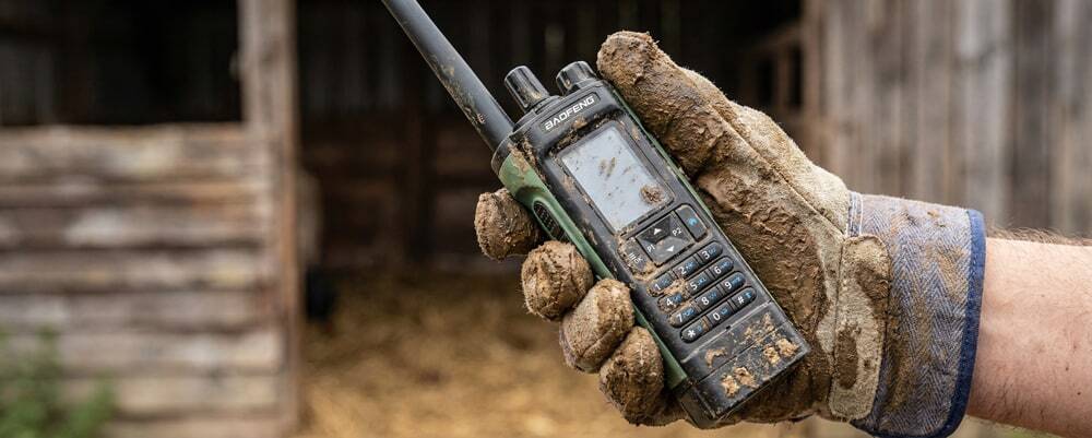 A close-up of a farmer with muddy gloves holding a durable agricultural walkie-talkie.