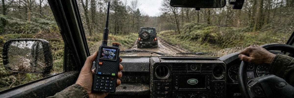 An off-road driver stands on a muddy forest trail, using a handheld radio to guide the convoy.