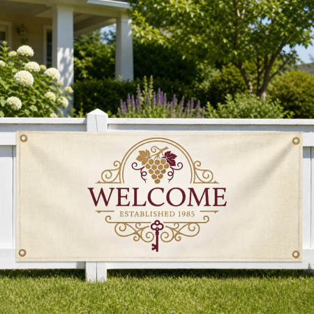A beige decorative “WELCOME” banner with gold scrollwork, grapes, and the text “Established 1985,” hanging on a white fence in a sunny garden with green shrubs and a house in the background.