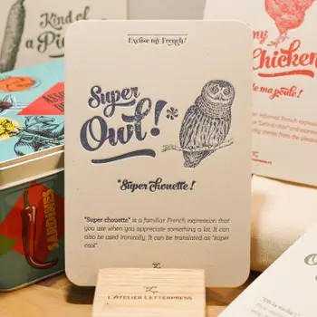 A "Chouette" card from L'Atelier Letterpress's "Excuse My French" series, displayed on a stand on a table. A sketch of a owl is at the top of the card. Below it in sweep cursive text reads, "Super Owl! Super chouette!" It is accompanied with an explanatory paragraph:
"'Super chouette' is a familiar French expression that you use when you appreciate something a lot. It can also be used ironically. It can be translated as "super cool."