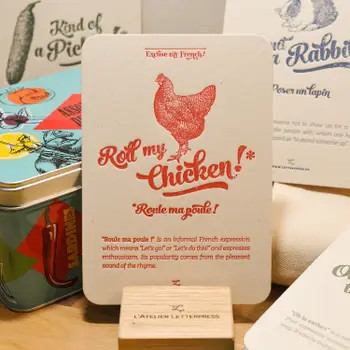 A "Poule" card from L'Atelier Letterpress's "Excuse My French" series, displayed on a stand on a table. A sketch of a chicken is at the top of the card. Below it in sweep cursive text reads, "Roll my chicken! Roule ma poule!" It is accompanied with an explanatory paragraph: "'Roule ma poule!' is an informal French expression which means "Let's go!" or "Let's do this!" and expresses enthusiasm. Its popularity comes from the pleasant sound of the rhyme.