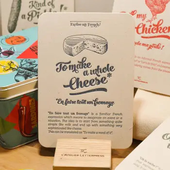 A "Fromage" card from L'Atelier Letterpress's "Excuse My French" series, displayed on a stand on a table. A sketch of cheese wheel is at the top of the card. Below it in sweep cursive text reads, "To make a whole cheese. En faire tout un fromage." It is accompanied with an explanatory paragraph: "'En faire tout un fromage' is a familiar French expression which means to exaggerate an event or a situation. The idea is to start from something quite simple like milk and up with something very sophisticated like cheese. This can be translated as, "to make a meal of it."