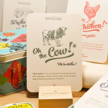 A "Vache" card from L'Atelier Letterpress's "Excuse My French" series, displayed on a stand on a table. A sketch of cow is at the top of the card. Below it in sweeping cursive reads, "Oh the cow! Oh la vache!" It is accompanied with an explanatory paragraph: "'Oh la vache!' is a very common French exclamation that expresses surprise, shock, often in an amused way. It can be translated by "Oh, damn!"