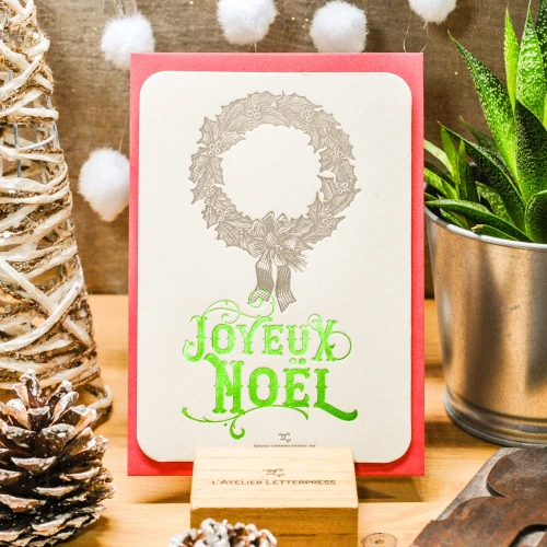 A greeting card that reads "Joyeux Noël" in bright green. Above it is an illustration of a wreath. The card sits on a stand.