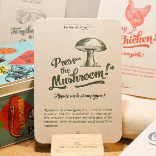 A "Champignon" card from L'Atelier Letterpress's "Excuse My French" series, displayed on a stand on a table. A sketch of a mushroom is at the top of the card. Below it in sweep cursive text reads,  "Press the mushroom! Appuie sur le champignon." It is accompanied with an explanatory paragraph: "'Appuie sur le champignon!' is a familiar French expression that can be translated as "Step on it!" This expression comes from the early days of the automobile, when the accelerator pedal looked like a mushroom."