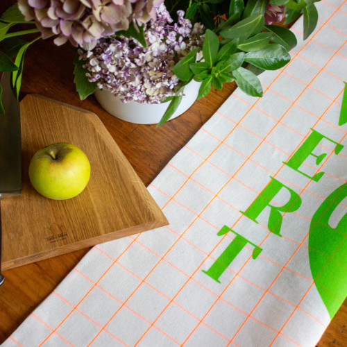 A close-up angle of the Reine Mère "Apple" tea towel. You can see a green apple on a white towel with a grid pattern. Beside it says "Vert" in bold green font. The towel is partly obscured by a cup holder holding an apple.