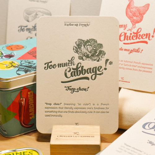 A "Chou" card from L'Atelier Letterpress's "Excuse My French" series, displayed on a stand on a table. A sketch of a cabbage is at the top of the card. Below it in sweep cursive text reads, "Too much cabbage! Trop chou!"It is accompanied with an explanatory paragraph: "'Trop chou!' (meaning "so cute!") is a French expression that literally expresses one's fondness for something that one finds absolutely cute. It can also be used ironically."