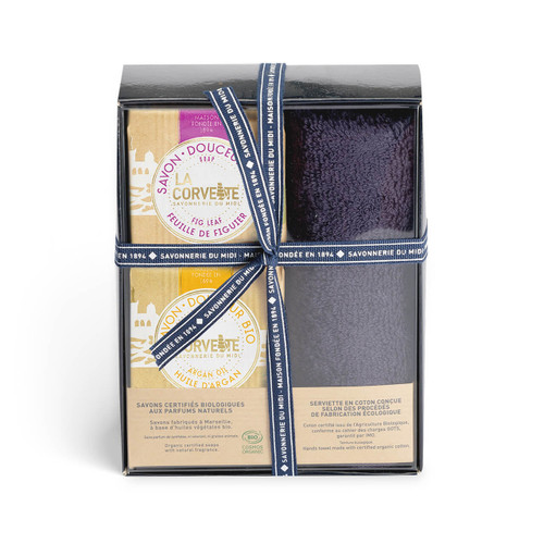 A clear "bath box" from La Corvette containing an assorted collection of toiletries. On the left are two organic soap bars, and on the right is gray cotton towel. The box is held together with a ribbon.