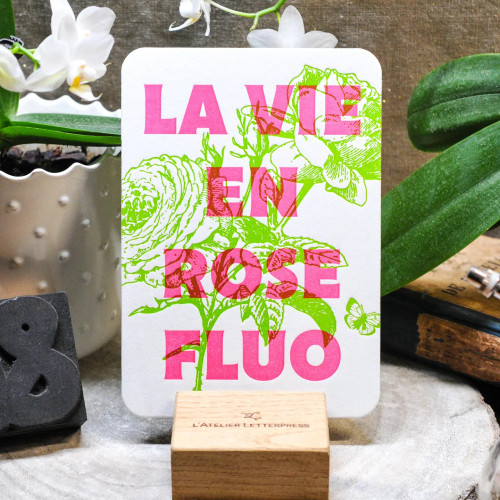 A card from L'Atelier Letterpress that reads, "La vie en rose fluo" in pink lettering in front of an illustration of a green rose. The card sits on a display stand.