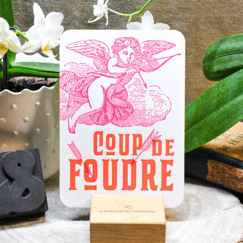 A card from L'Atelier Letterpress that reads, "Coup de foudre" in red lettering in front of an illustration of baby Cupid drawing a bow. The card sits on a display stand.
