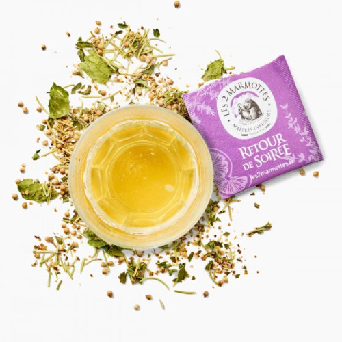 A white teacup holding a serving of Les 2 Marmottes's "Retour de Soirée" herbal tea, a warm yellow brew, surrounded by dried herbs.