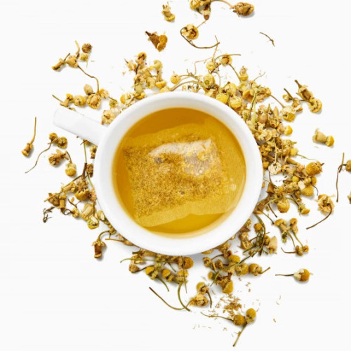 A white teacup holding a serving of Les 2 Marmottes's chamomile herbal tea, a warm yellow brew, surrounded by dried chamomile flowers.