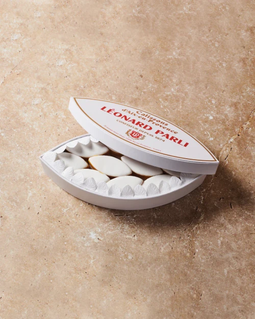 An oblong tin containing 230 grams of Provence calissons from Léonard Parli. The tin is white with red lettering that says, "Calissons d'Aix-en-Provence: Léonard Parli, Confiseur depuis 1874." The tin sits on granite countertop.