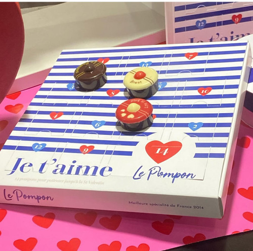 A Valentine's Calendar from Le Pompon containing 150 grams of gourmet chocolate bonbons, displayed on a pink tablecloth. The bonbons are shaped in homage to the French navy's sailor cap, cylindrical with a "pompon" on top. The box has many sealed flaps which contain chocolate, similar to an Advent calendar. The calendar is striped blue and white.