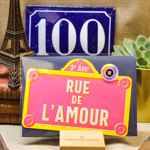 A card that reads "3e Arrondissements: Rue de l'Amour," hand printed in the style of a pink street sign, from L'Atelier Letterpress. The card is displayed on a stand.