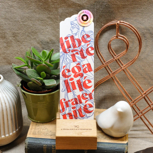 A hand printed L'Atelier Letterpress bookmare which reads, "Liberté, Égalité, Fraternité," the national motto of France, in front of an illustration of Marianne, the personification of the French Republic. The bookmark is displayed prominently on a bookshelf.