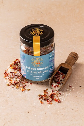 A jar containing 90 grams of salt with tomatoes and olives from Comptoir & Co. The jar sits on a stone surface next to a salt scoop.