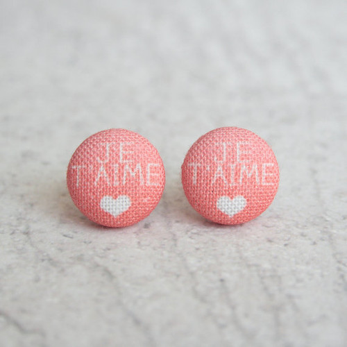 A pair of pink fabric button earrings with white text that says, "Je t'aime," with a white heart beneath it, from Rachel O's. The earrings are on a gray stone counter top.